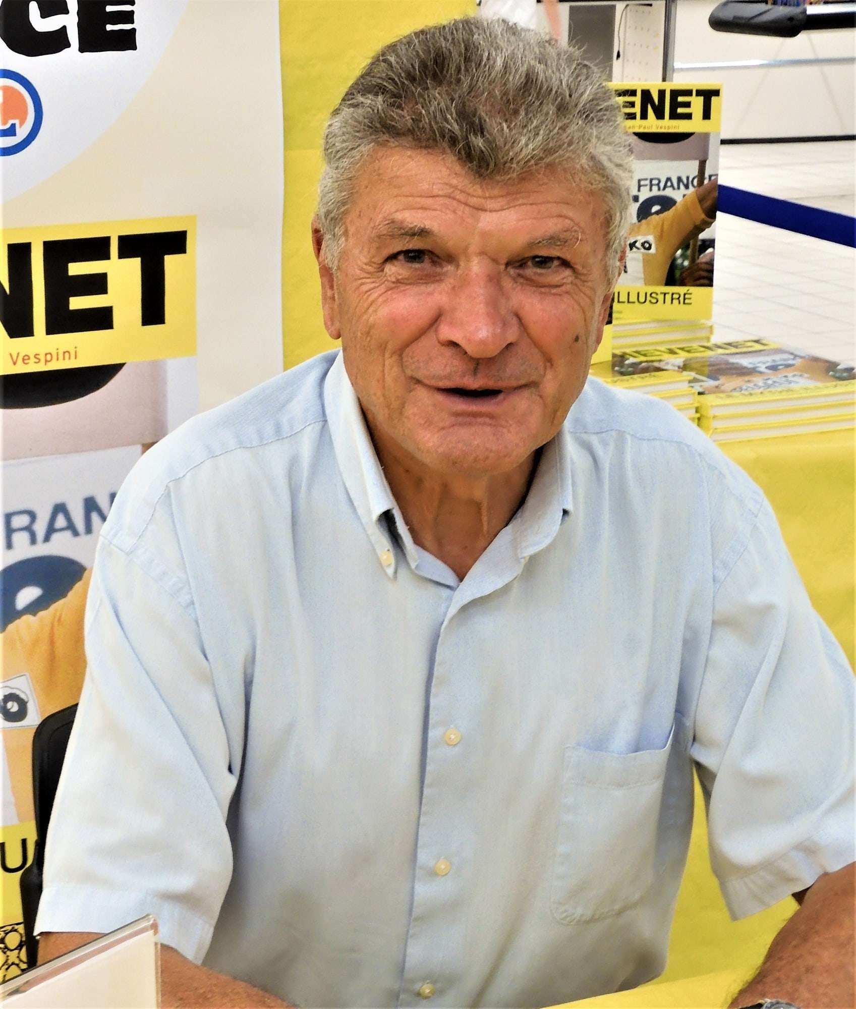 Bernard Thévenet assis à une table de dédicace devant des affiches jaunes