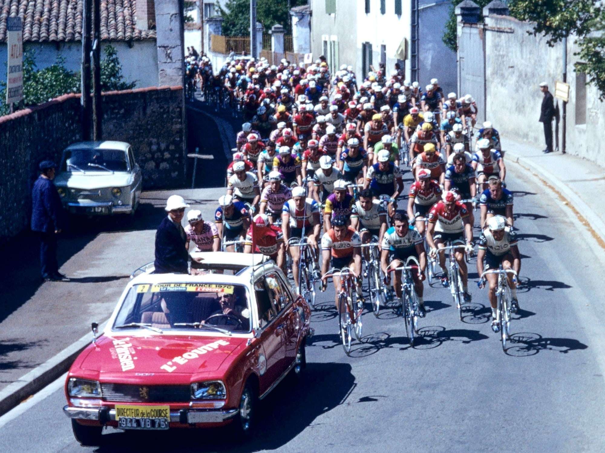 Le peloton du Tour de France 1970 dans une rue de village, précédé d'une voiture rouge